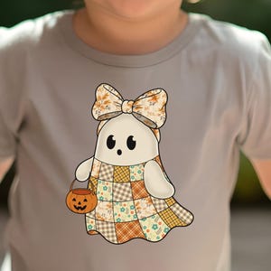 Pode incluir: Camiseta cinza claro com um desenho de fantasma de desenho animado. O fantasma usa um la&ccedil;o e um vestido de retalhos e carrega um balde de ab&oacute;bora. O la&ccedil;o e o vestido apresentam padr&otilde;es florais e xadrez. O fantasma tem olhos pretos e uma express&atilde;o amig&aacute;vel.