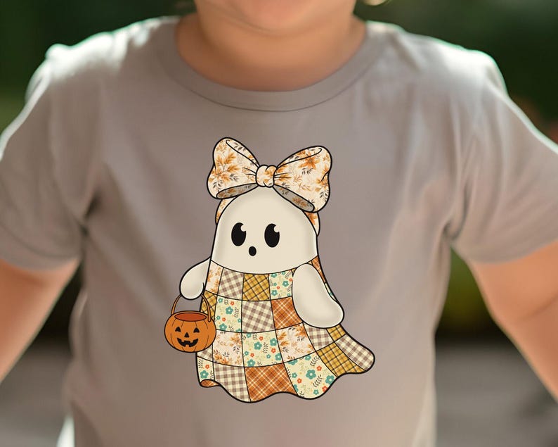 Pode incluir: Camiseta cinza claro com um desenho de fantasma de desenho animado. O fantasma usa um la&ccedil;o e um vestido de retalhos e carrega um balde de ab&oacute;bora. O la&ccedil;o e o vestido apresentam padr&otilde;es florais e xadrez. O fantasma tem olhos pretos e uma express&atilde;o amig&aacute;vel.