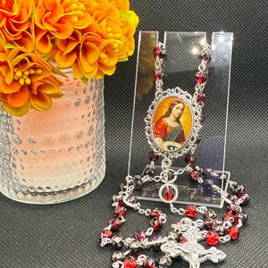 May include: A rosary with red and black faceted beads and a silver-toned crucifix. The centerpiece features an oval image of a saint. A vase of orange flowers sits to the left of the rosary.