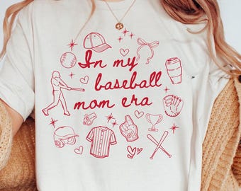Op de afbeelding: Een ivoorwit T-shirt met een rood grafisch ontwerp met de tekst "In my baseball mom era" en baseball-thema illustraties zoals een honkbal, knuppel, handschoen en een shirt.