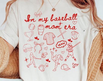 Op de afbeelding: Een wit T-shirt met een rode afbeelding waarop "In my baseball mom era" staat en baseball-thema illustraties.