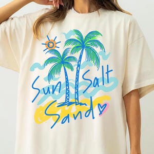 Könnte beinhalten: Elfenbeinfarbenes T-Shirt mit einem Strandmotiv. Es zeigt Palmen, eine Sonne und Wellen mit den Worten "Sun Salt Sandy" in blauer Schrift. Das Design enthält gelbe, blaue und grüne Akzente. Das T-Shirt hat eine lockere Passform.
