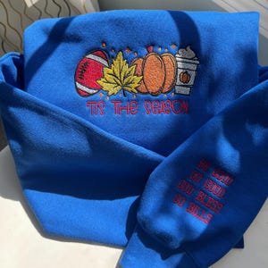 May include: A royal blue sweatshirt with embroidered fall-themed designs. The design includes a football, pumpkins, and a coffee cup, with the phrase "Tis the Season." The sleeve has the embroidered text "Be Good, Do Good, God Bless, Go Bills."