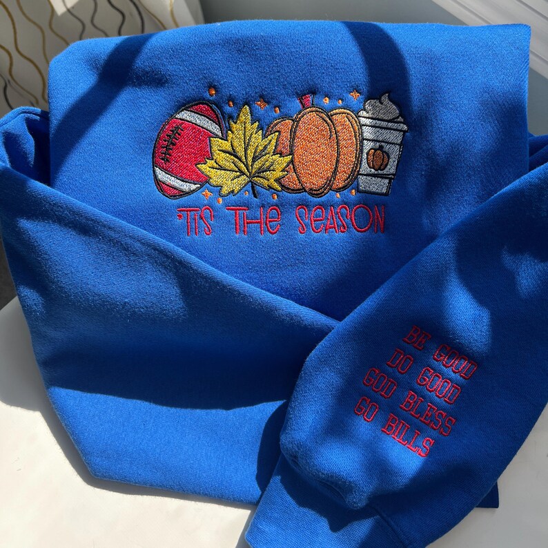 May include: A royal blue sweatshirt with embroidered fall-themed designs. The design includes a football, pumpkins, and a coffee cup, with the phrase "Tis the Season." The sleeve has the embroidered text "Be Good, Do Good, God Bless, Go Bills."