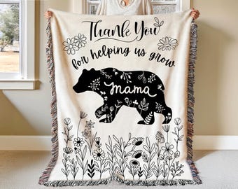 May include: A cream-colored woven throw blanket with a black bear silhouette and the text "Thank You for helping us grow mama" surrounded by floral designs.