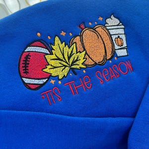 May include: A royal blue sweatshirt with an embroidered design featuring a football, pumpkins, a maple leaf, and a coffee cup. The text "Tis The Season" is embroidered in red. The design has a fall theme.