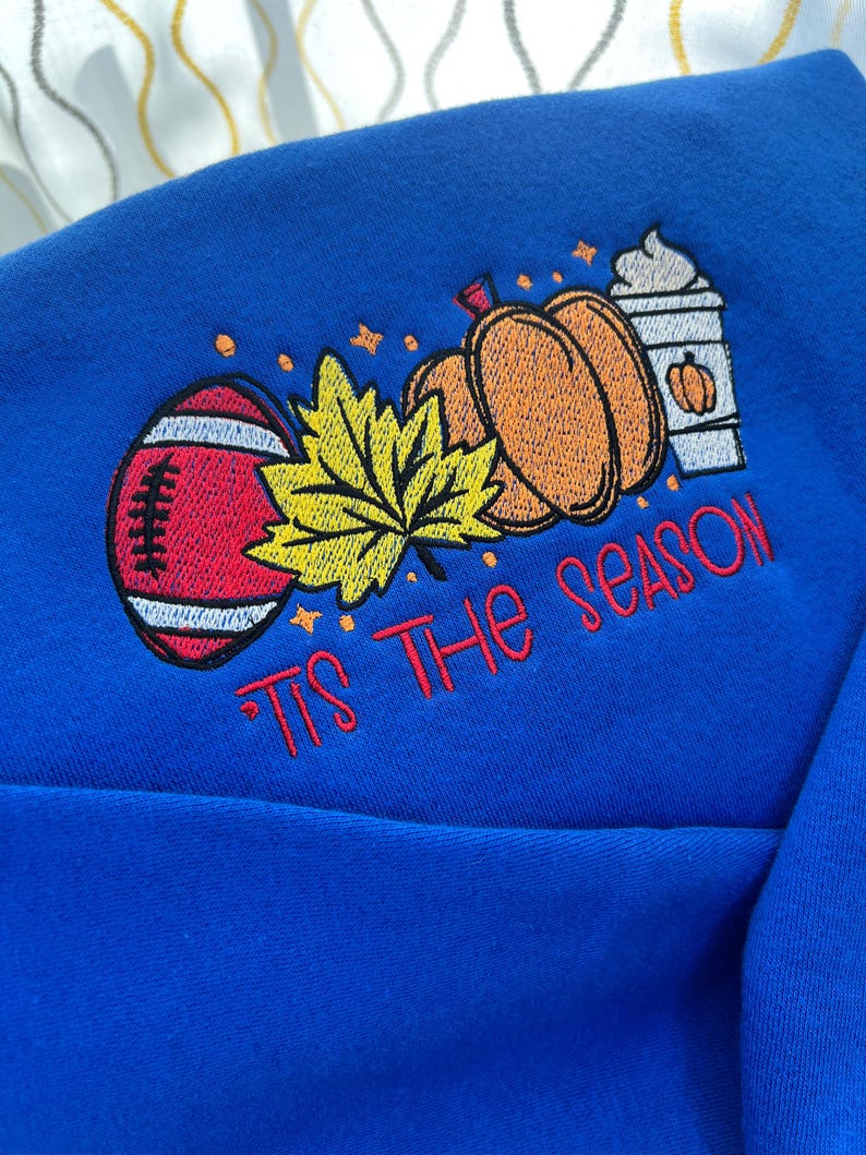 May include: A royal blue sweatshirt with an embroidered design featuring a football, pumpkins, a maple leaf, and a coffee cup. The text "Tis The Season" is embroidered in red. The design has a fall theme.
