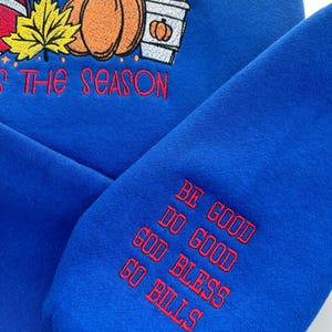May include: Royal blue sweatshirt with embroidered fall-themed design. Features pumpkins, a leaf, and a coffee cup with the text "'Tis the Season". The sleeve has the text "Be Good, Do Good, God Bless, Go Bills".