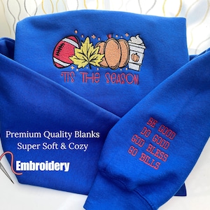 May include: A royal blue sweatshirt with embroidered fall-themed graphics, including a football, pumpkins, and a coffee cup. The text "'Tis the Season" is embroidered in red. The sleeve features the text "Be Good, Do Good, God Bless, Go Bills".