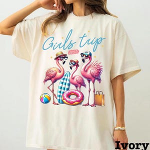 May include: Ivory-colored t-shirt featuring a graphic of three pink flamingos wearing sunglasses and hats, with a surfboard, donut float, and beach ball. The text "Girls trip 2025" is written in blue script.