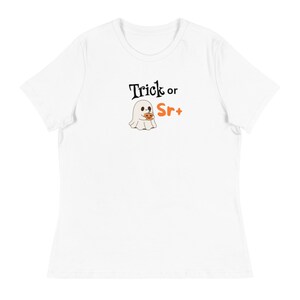 Puede incluir: Camiseta blanca con cuello redondo con un diseño temático de Halloween. El diseño incluye el texto "Trick or Sr+" encima de un fantasma de dibujos animados sosteniendo un dulce con forma de calabaza. El fantasma es blanco y la calabaza es naranja.