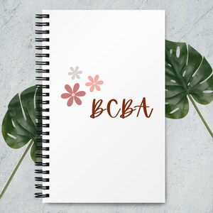 以下が含まれることがあります： 茶色の筆記体で「BCBA」の文字が入った、白いリング綴じノート。 ノートの表紙には、ピンクとグレーの色合いの3つの花の模様があります。 ノートは緑の葉を背景に配置されています。