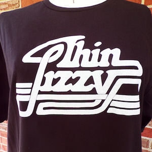 May include: Black t-shirt with a white "Thin Lizzy" logo printed on the front. The logo is in a retro style with white stripes.