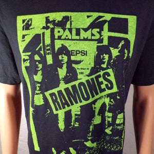 May include: Black t-shirt with a green graphic print of the Ramones band. The print features the band members in front of a building with the words "Palms" and "Pepsi" visible. The text "The Return of The Ramones" is also included in the print.
