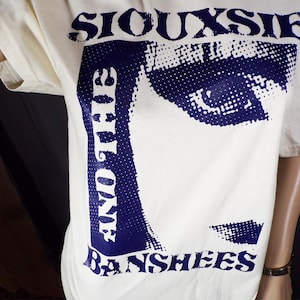 May include: A white t-shirt with a blue graphic print of Siouxsie and the Banshees. The graphic features a close-up of a face with a dotted pattern.