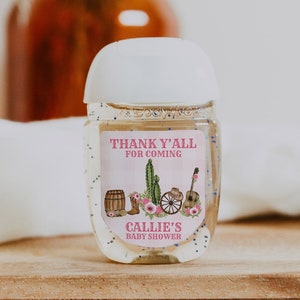 May include: A clear plastic bottle of hand sanitizer with a white cap and a pink and white label that says "Thank y'all for coming Callie's Baby Shower". The label also features a cactus, cowboy boots, a wagon wheel, and a guitar.