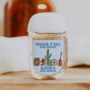 May include: A clear plastic bottle of hand sanitiser with a white pump top and a blue and white label that says "Thank Y'all for Coming Katie's Baby Shower". The label has a cactus, a cowboy boot, a wagon wheel, and a guitar on it.
