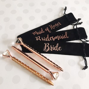 May include: Three rose gold pens with diamond-shaped tops, each in a black velvet pouch with gold lettering. The pouches read "Maid of Honor", "Bridesmaid", and "Bride".