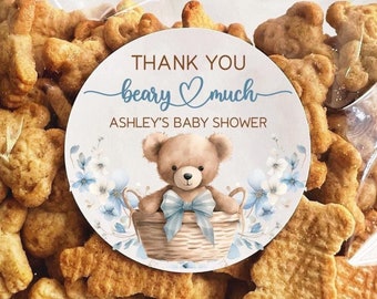 May include: A clear plastic bag filled with bear-shaped crackers. A round sticker with a blue and white floral design and a teddy bear holding a blue bow is attached to the bag. The sticker says "Thank you beary much Ashley's Baby Shower".