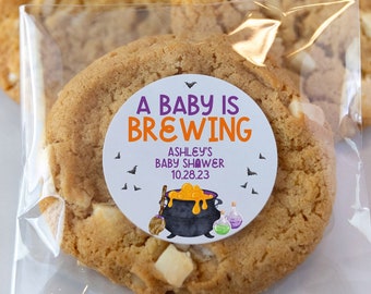 May include: A clear plastic bag filled with three sugar cookies. The cookie in the front has a round sticker with a purple and orange design that says "A Baby is Brewing" with the text "Ashley's Baby Shower 10.28.23".