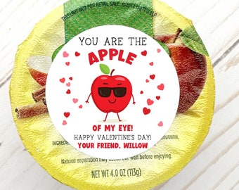 May include: A red apple with sunglasses and a smile on a white circle with the text "You are the apple of my eye! Happy Valentine's Day! Your friend, Willow" and pink hearts. The circle is on top of a yellow container with a red apple on the side.
