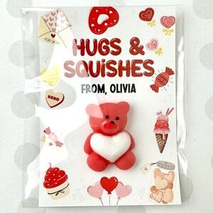 May include: A red and white Valentine's Day card with a red teddy bear holding a white heart. The card says "Hugs & Squishes" and "From, Olivia".