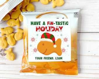 May include: A snack bag with a holiday-themed label featuring a cartoon goldfish wearing a Santa hat. The text reads "Have a Fin-tastic Holiday" and "Your Friend, Liam." Goldfish crackers are scattered around the bag on a white wooden surface.