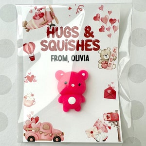 May include: A Valentine's Day card with a pink and red bear-shaped squishy toy. The card reads "Hugs & Squishes From, Olivia" and features illustrations of hearts, a pink truck, and a gnome holding a letter.
