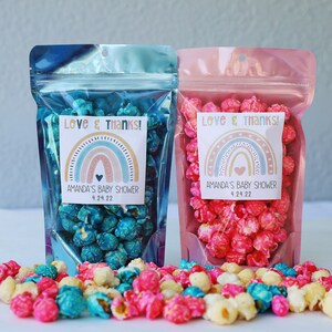 May include: Two clear plastic bags filled with popcorn. The bag on the left is blue and the bag on the right is pink. Both bags have a sticker with a rainbow design and the text "Love & THANKS!" and "Amanda's Baby Shower 4.24.22".