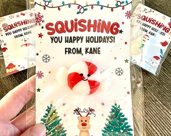 Pode incluir: Um cartão branco com decorações de Natal vermelhas e verdes e o texto "SQUISHING YOU HAPPY HOLIDAYS! FROM, KANE". Um bastão de caramelo branco com uma faixa vermelha e um rosto sorridente está preso ao cartão.