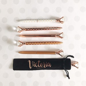 May include: Set of five gold pens with white polka dots and diamond-shaped tops. The pens are in a black velvet pouch with the name "Victoria" written in gold lettering.
