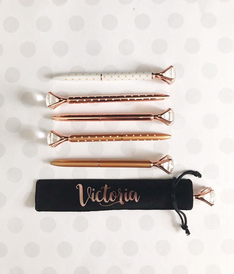May include: Set of five gold pens with white polka dots and diamond-shaped tops. The pens are in a black velvet pouch with the name "Victoria" written in gold lettering.