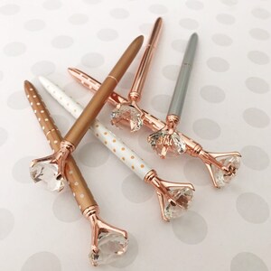 May include: Five pens with rose gold accents and clear rhinestone tops. The pens have white and gold polka dot patterns and solid rose gold.