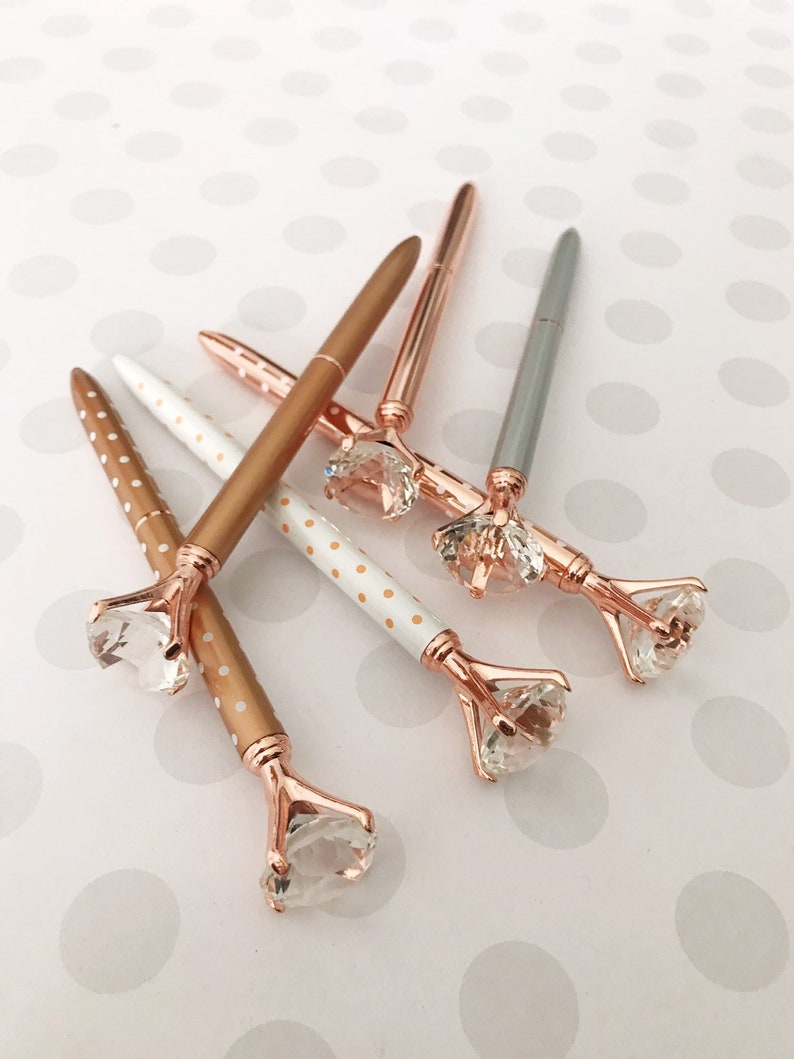 May include: Five pens with rose gold accents and clear rhinestone tops. The pens have white and gold polka dot patterns and solid rose gold.