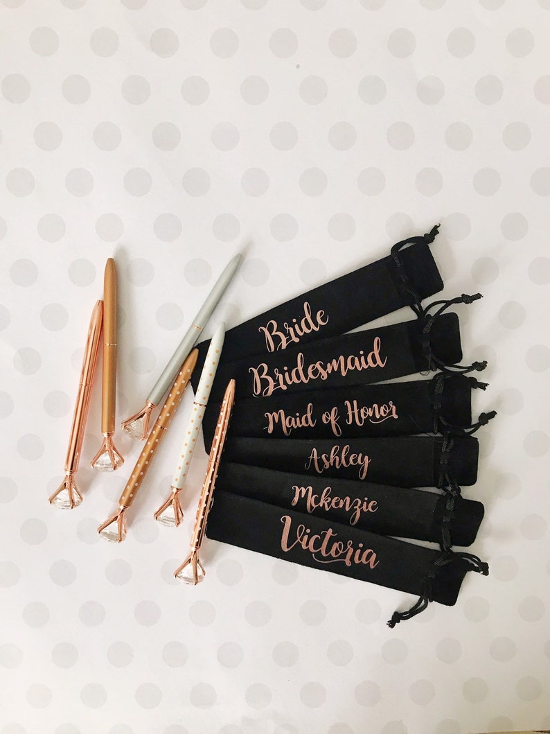 May include: Six black velvet pouches with gold lettering, each with a different title: Bride, Bridesmaid, Maid of Honor, Ashley, McKenzie, Victoria. Each pouch contains a rose gold pen with a clear rhinestone on the end.