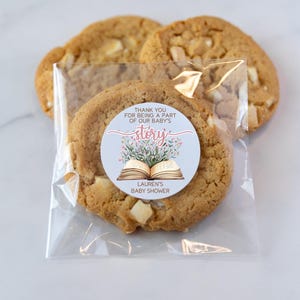May include: A clear plastic bag filled with cookies. A round sticker with a floral design and an open book is attached to one of the cookies. The sticker says "Thank you for being a part of our baby's story. Lauren's Baby Shower."