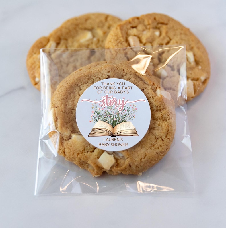 May include: A clear plastic bag filled with cookies. A round sticker with a floral design and an open book is attached to one of the cookies. The sticker says "Thank you for being a part of our baby's story. Lauren's Baby Shower."