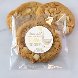 May include: A clear plastic bag with a white and yellow sticker that says "Thanks for celebrating our Silly Goose Emma's 1st Birthday". The sticker has a cartoon goose with a yellow balloon. The bag contains two cookies with white chocolate chips.