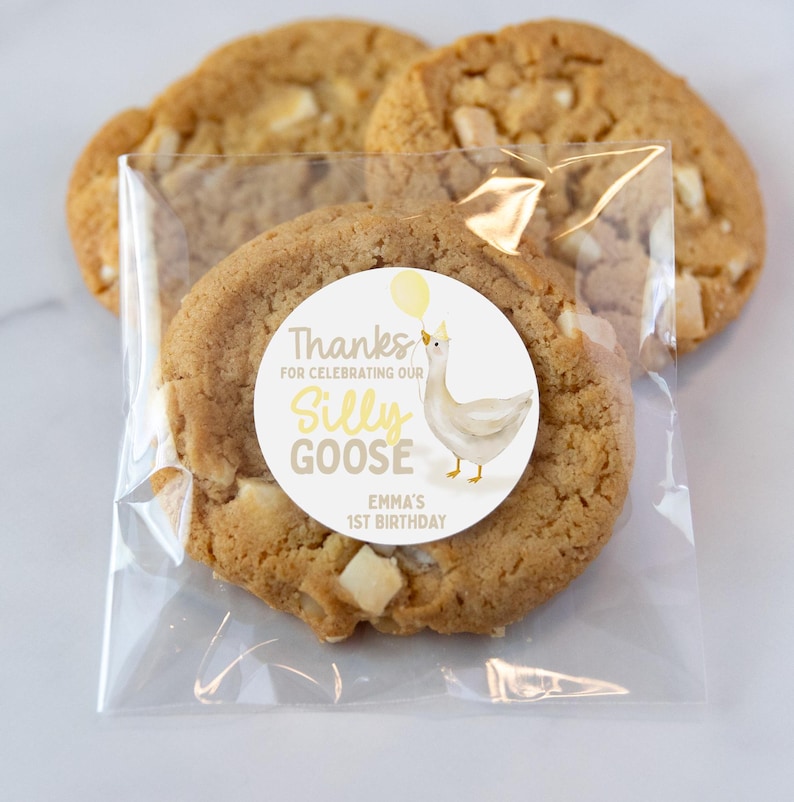 May include: A clear plastic bag with a white and yellow sticker that says "Thanks for celebrating our Silly Goose Emma's 1st Birthday". The sticker has a cartoon goose with a yellow balloon. The bag contains two cookies with white chocolate chips.
