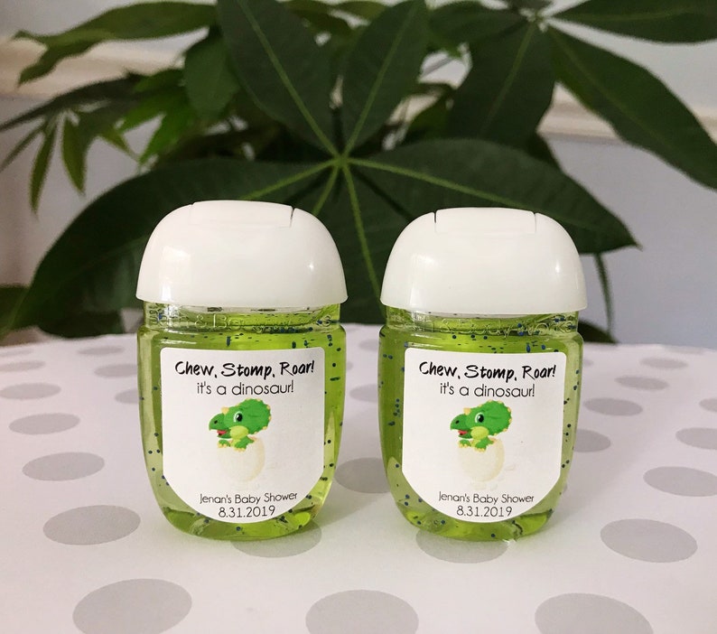 May include: Two green hand sanitiser bottles with white lids and labels that say "Chew, Stomp, Roar! It's a dinosaur!" and "Jenan's Baby Shower 8.31.2019".