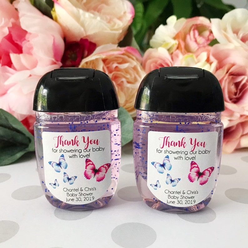 Puede incluir: Dos peque&ntilde;as botellas transparentes de gel desinfectante para manos con etiquetas que dicen "&iexcl;Gracias por consentir a nuestro beb&eacute; con amor!" Las etiquetas tambi&eacute;n incluyen mariposas y el texto "Chantel & Chris's Baby Shower June 30, 2019".