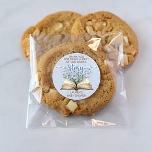 May include: A blue and white book with flowers on the cover. The book is open and the text "Thank you for being a part of our baby's story" is visible. The sticker is on a clear plastic bag with cookies inside. The text "Lauren's Baby Shower" is also visible on the sticker.