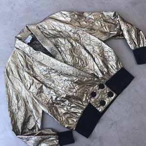 May include: A gold metallic wrap top with long sleeves and black ribbed cuffs. The top features a V-neck and a black band with four decorative buttons at the waist. The fabric has a textured, crinkled appearance.