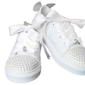 May include: White lace sneakers with a satin ribbon bow and pearl embellishments. The sneakers have small angel wings attached to the back.