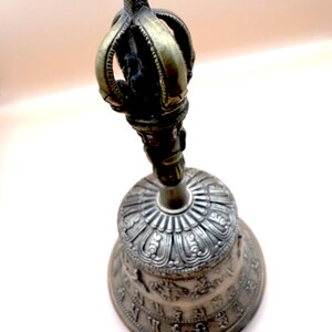 May include: A silver and gold bell with intricate carvings and a crown-shaped handle. The bell is decorated with a circular pattern and symbols.