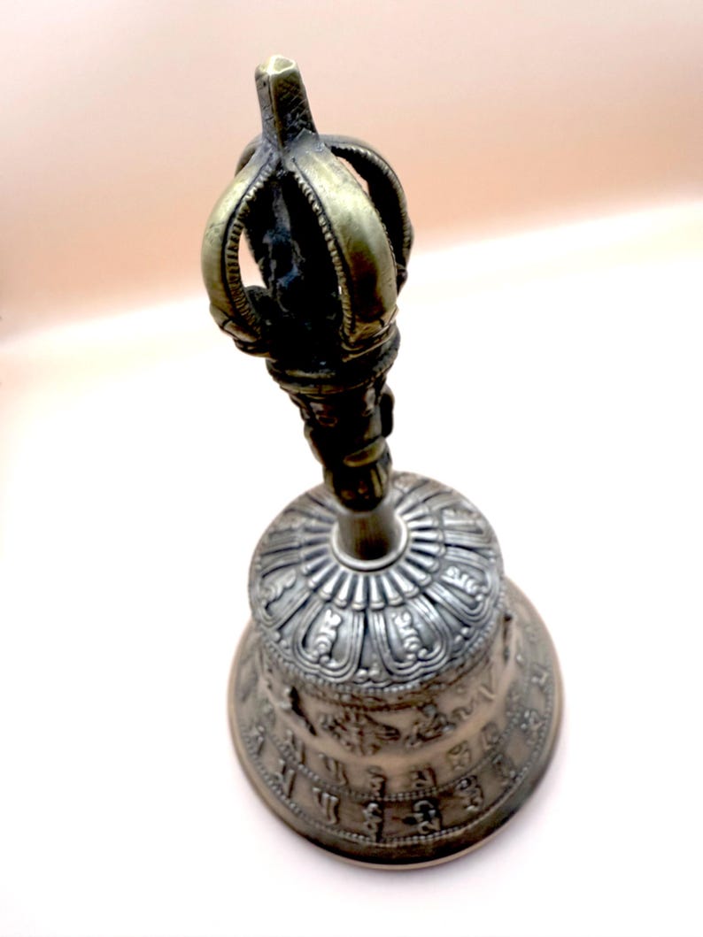 May include: A silver and gold bell with intricate carvings and a crown-shaped handle. The bell is decorated with a circular pattern and symbols.