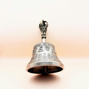 May include: A silver bell with a decorative handle, featuring intricate carvings and a design of seated figures. The bell is used in Tibetan Buddhism for meditation and rituals.
