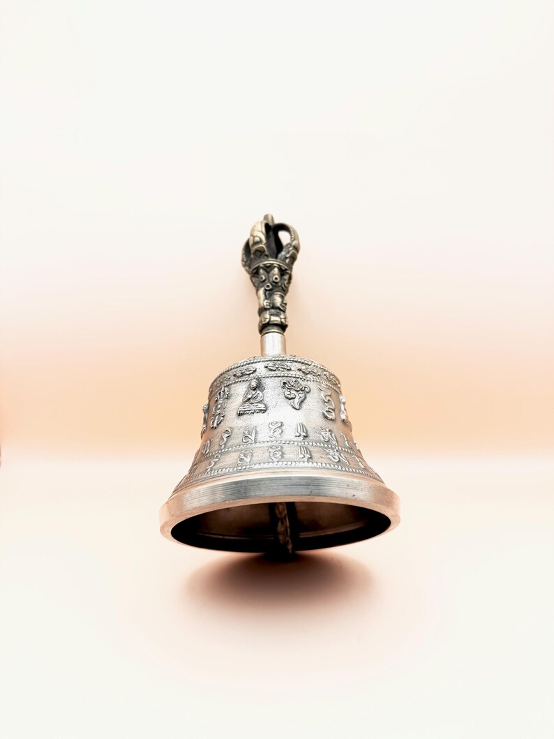 May include: A silver bell with a decorative handle, featuring intricate carvings and a design of seated figures. The bell is used in Tibetan Buddhism for meditation and rituals.