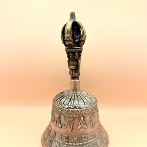May include: A silver bell with intricate carvings and a crown-shaped handle. The bell is decorated with Buddhist symbols and figures.