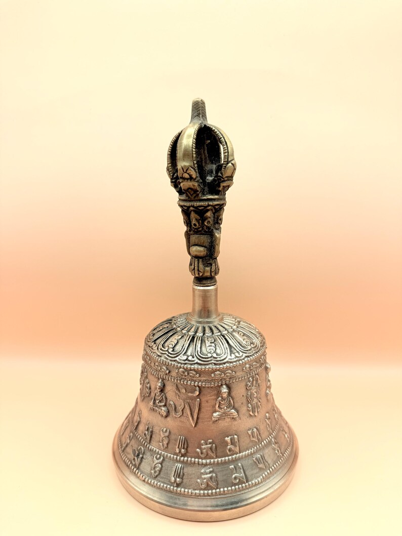 May include: A silver bell with intricate carvings and a crown-shaped handle. The bell is decorated with Buddhist symbols and figures.